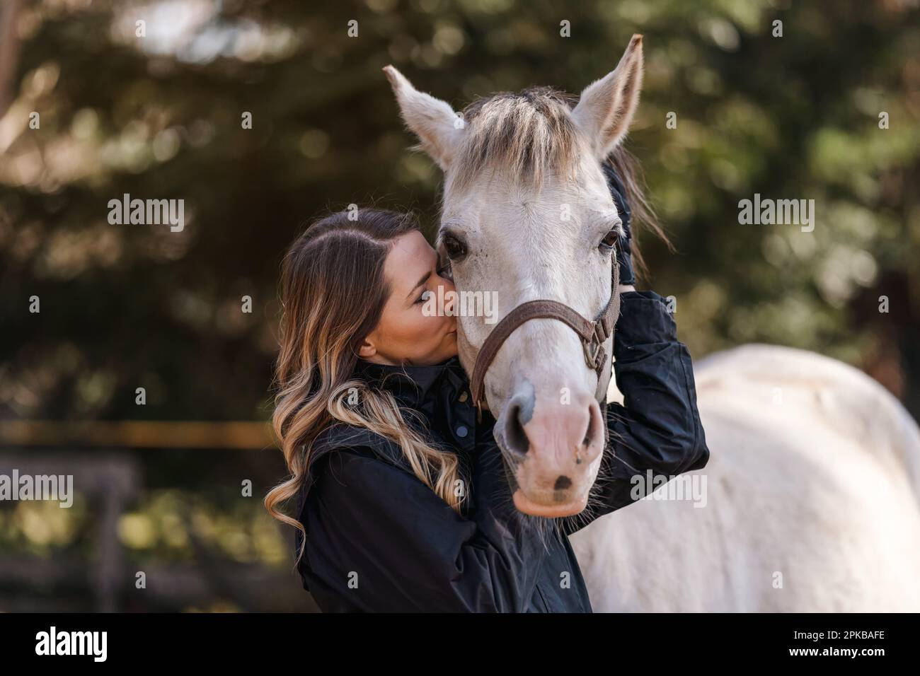 Jeune femme debout à côté des yeux blancs de cheval arabe fermé comme si elle s'embrasse ou sentait, brouillé arbre arrière-plan, gros plan Banque D'Images