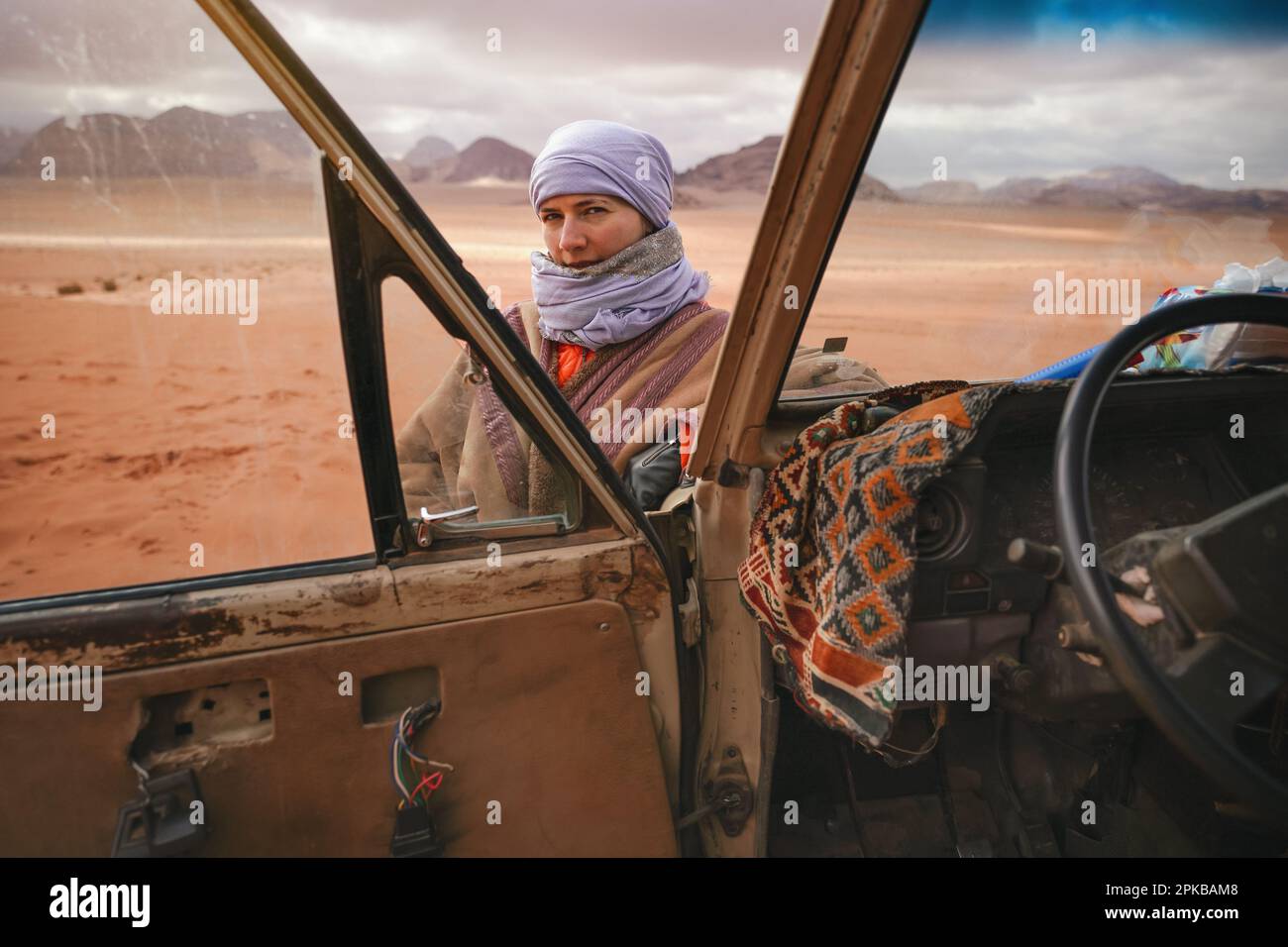 Jeune femme en manteau bédouin traditionnel - bisht - et foulard, posant à côté de l'ancien véhicule 4WD, regardant par-dessus la porte ouverte - paysage désertique de Wadi R Banque D'Images