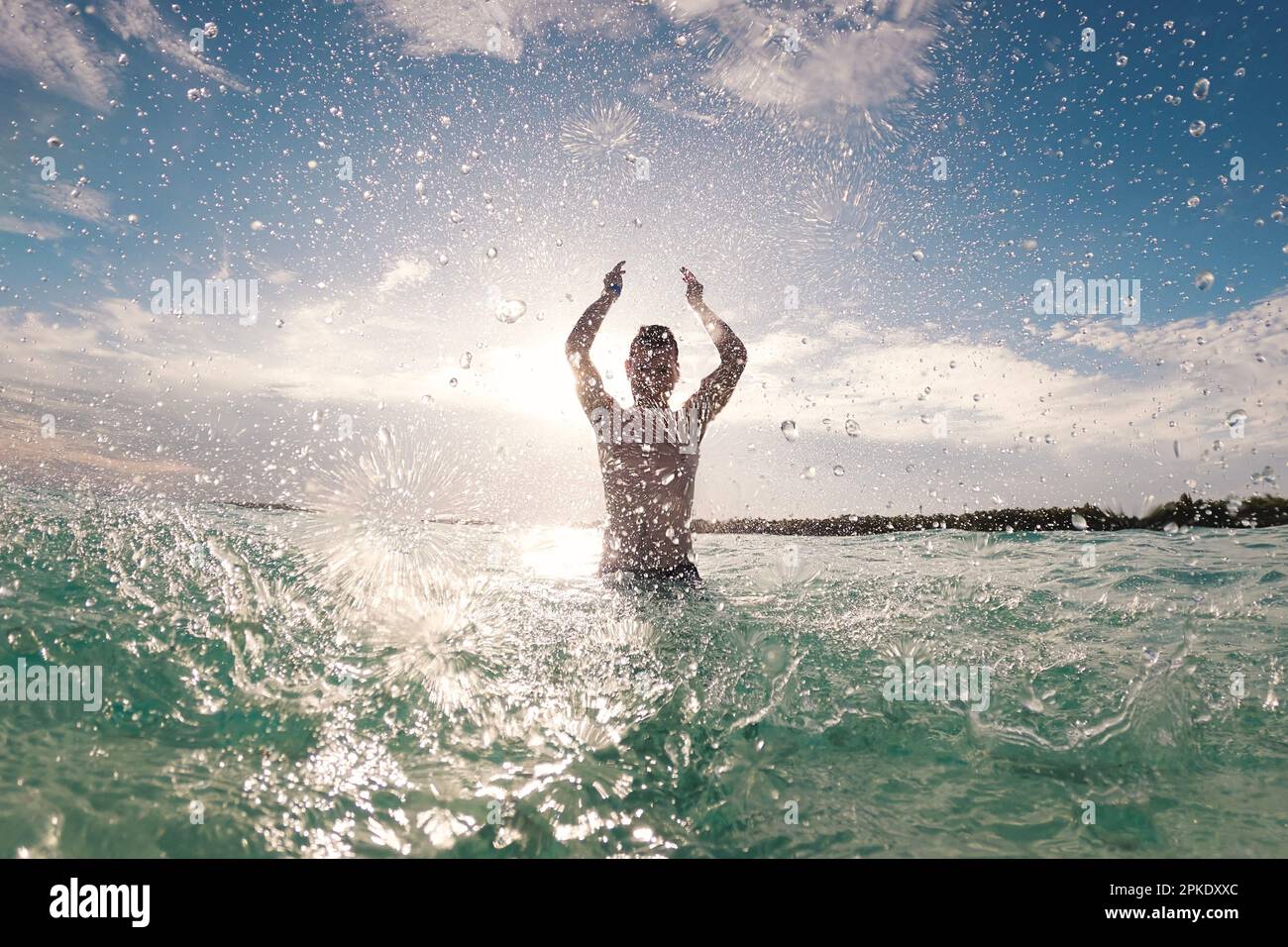 Silhouette d'homme joyeux debout en mer et éclaboussant l'eau vers l'appareil photo. Journée ensoleillée en destination tropicale pendant les vacances à la plage. Banque D'Images