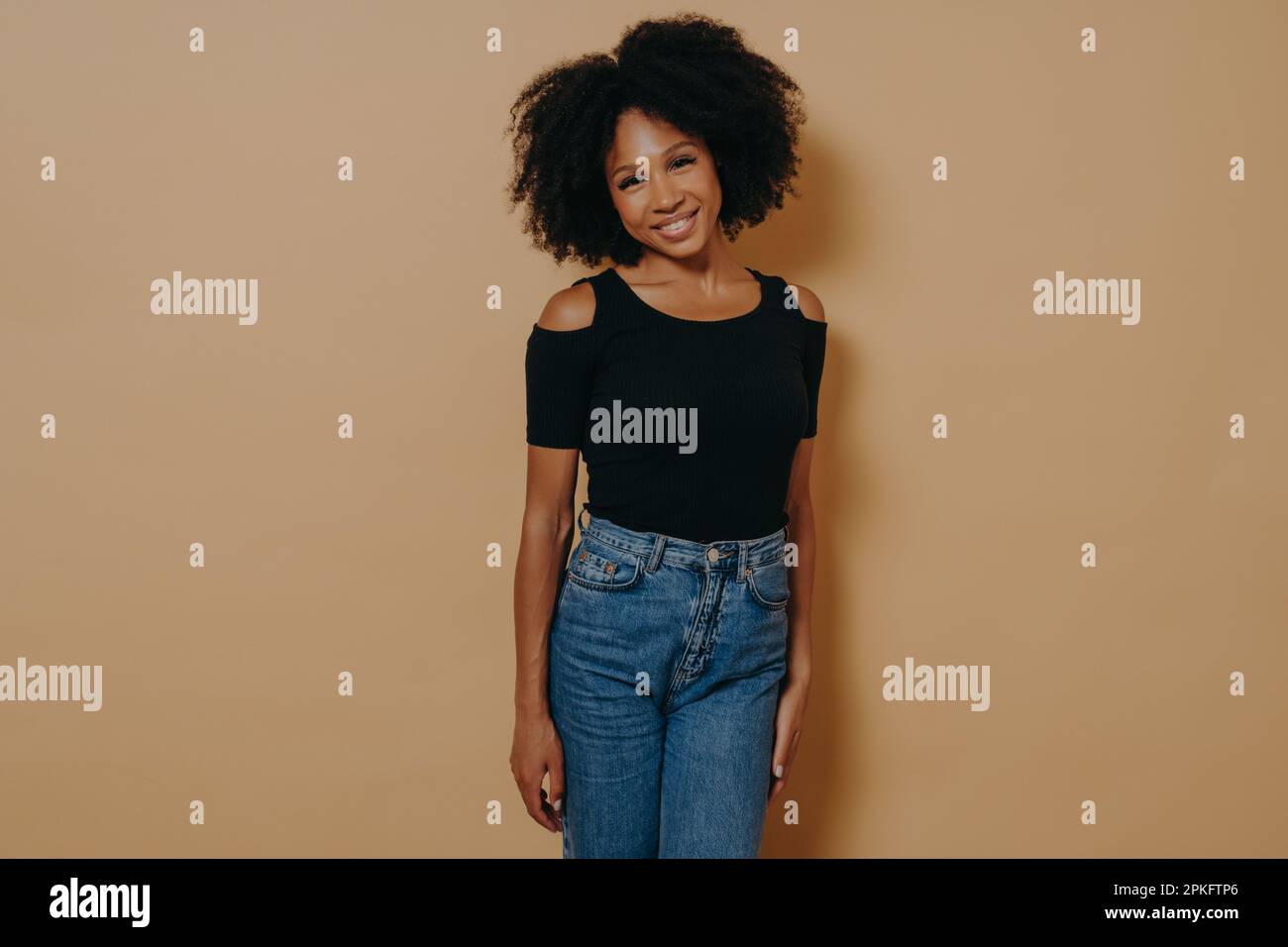 Photo d'intérieur de la jolie femme afro-américaine souriante dans une tenue décontractée regardant l'appareil photo avec un sourire timide et charmant, posant isolée sur le dos beige foncé Banque D'Images