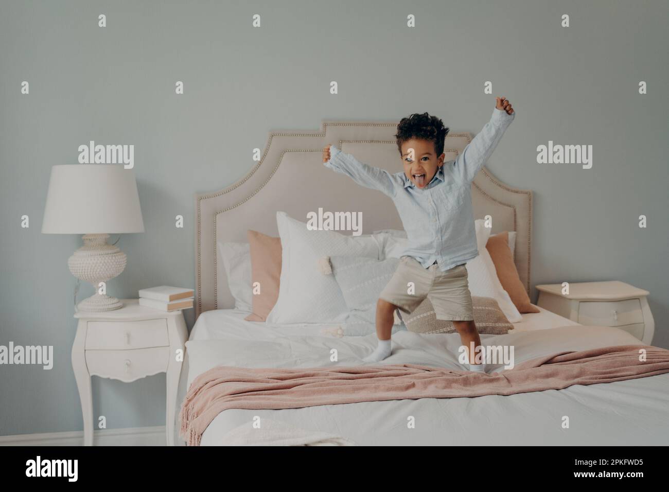 Adorable enfant africain mignon sautant sur le lit recouvert de couvre-lits dans des tons rose clair et blanc à côté de la lampe sur la table de chevet, enfant de race active mixte Banque D'Images