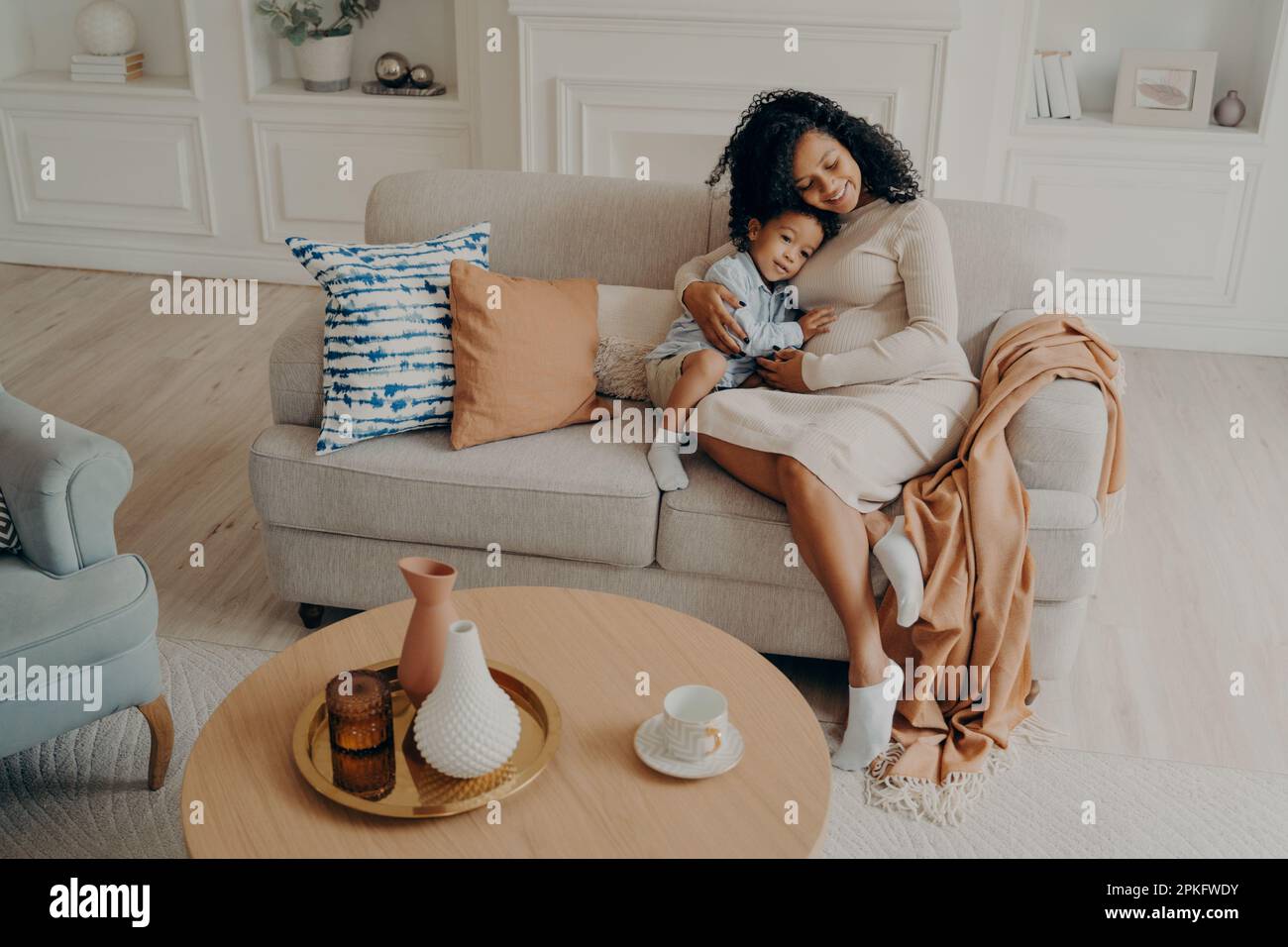 Maman enceinte africaine affectueuse embrassant le petit fils tout en étant assis sur un canapé confortable à côté de la table basse dans le salon, passant du temps de loisirs ensemble Banque D'Images