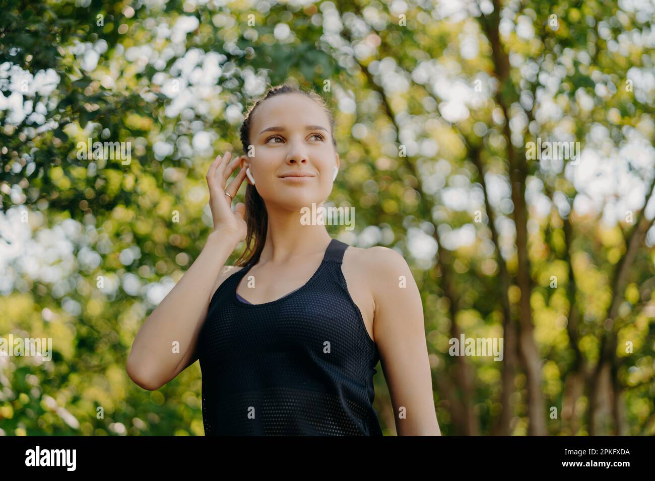 Prise de vue horizontale d'une jeune femme satisfaite tient la main sur l'écouteur écoute de musique préférée regarde loin a satisfait expression du visage vêtu de vêtements actifs e Banque D'Images