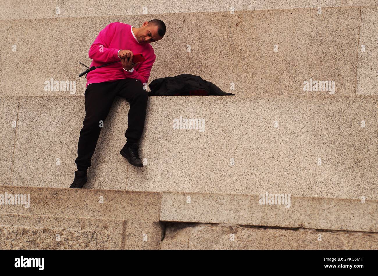 Un jeune asiatique assis sur la plinthe de la colonne de Nelson, Trafalgar Square, Londres. Royaume-Uni regardant son téléphone portant un pull rouge et un pantalon noir Banque D'Images