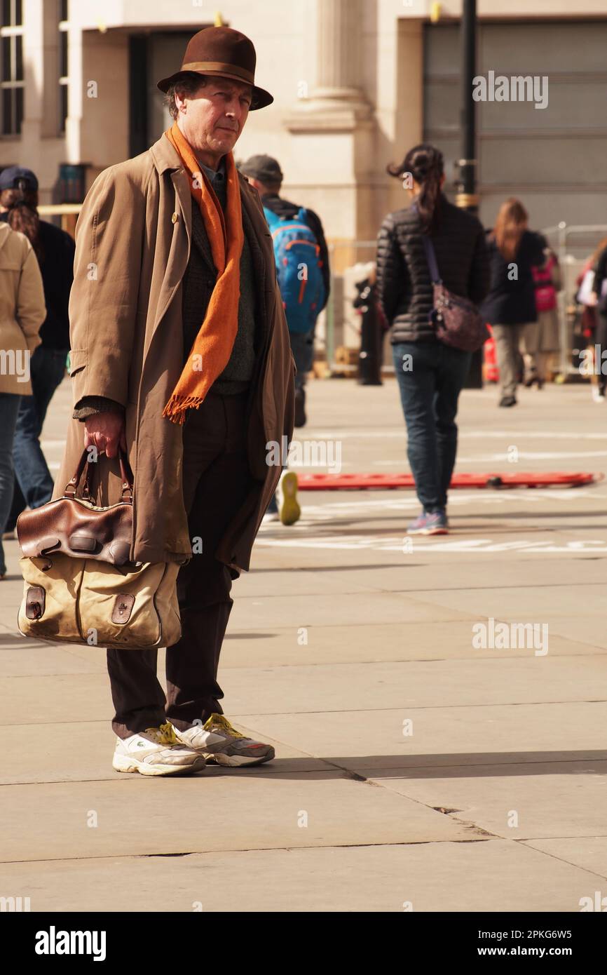 Un homme plus âgé de 60 ans et plus debout avec un long manteau, un grand sac, une écharpe orange, un chapeau marron debout à Trafalgar Square, Londres regardant et écoutant un bucker Banque D'Images