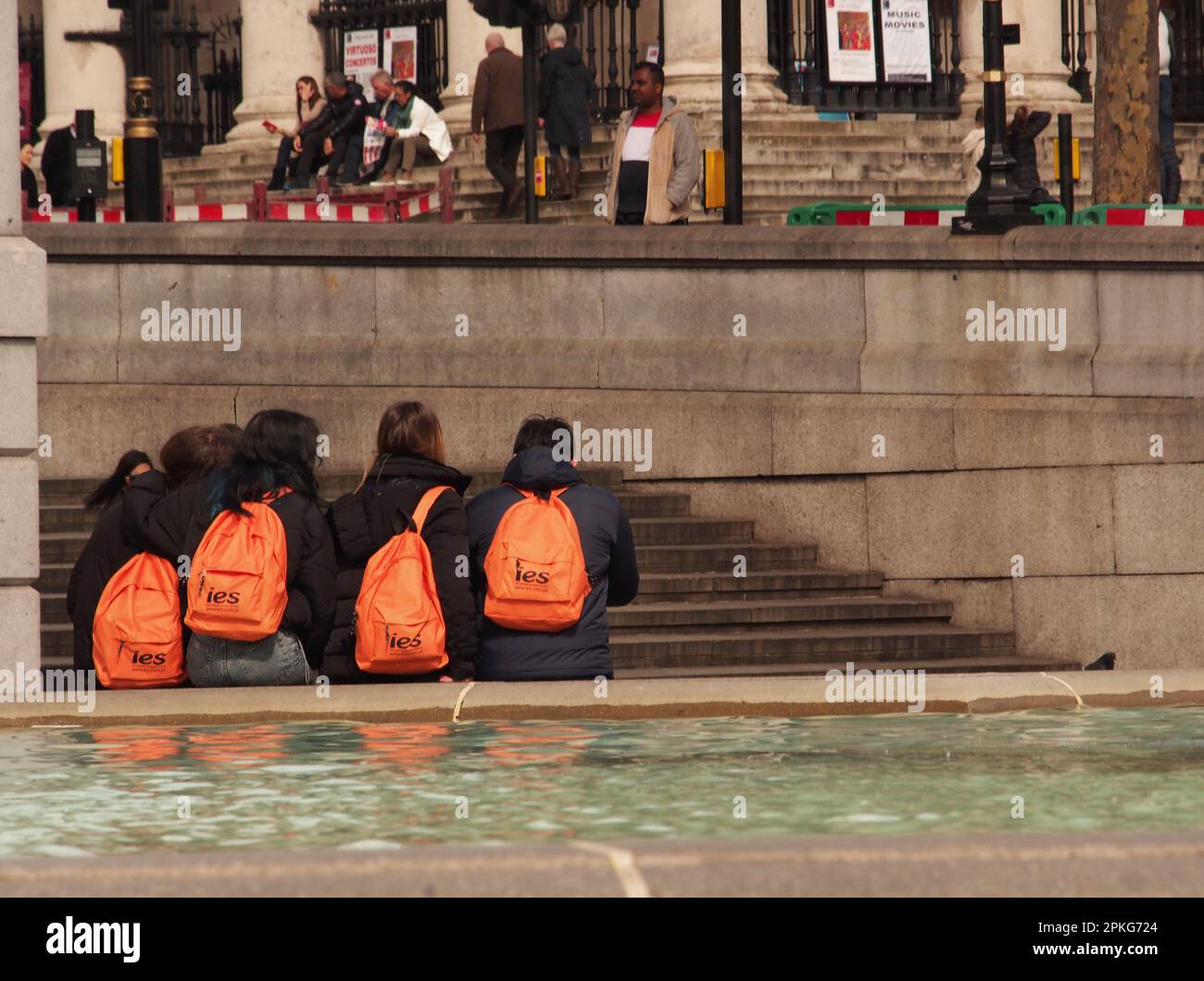 Quatre jeunes adolescents, du Portugal, visitant Trafalgar Square, Londres. Royaume-Uni assis sur le bord d'une piscine avec des sacs d'orange sur le dos Banque D'Images