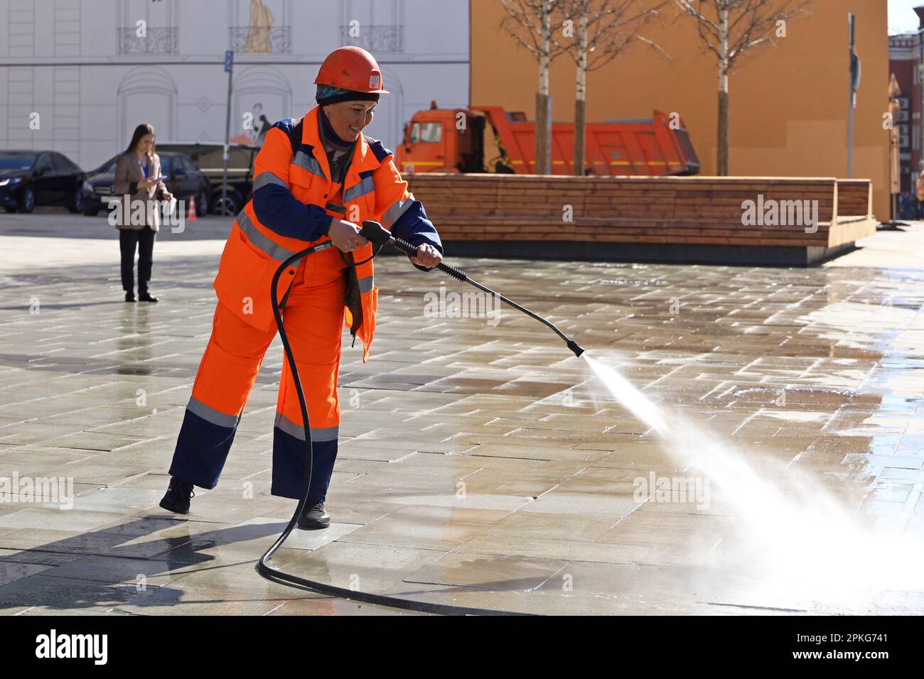 Bonne femme asiatique en uniforme de travailleur arroser le trottoir avec un tuyau. Nettoyage et désinfection des rues au printemps Banque D'Images