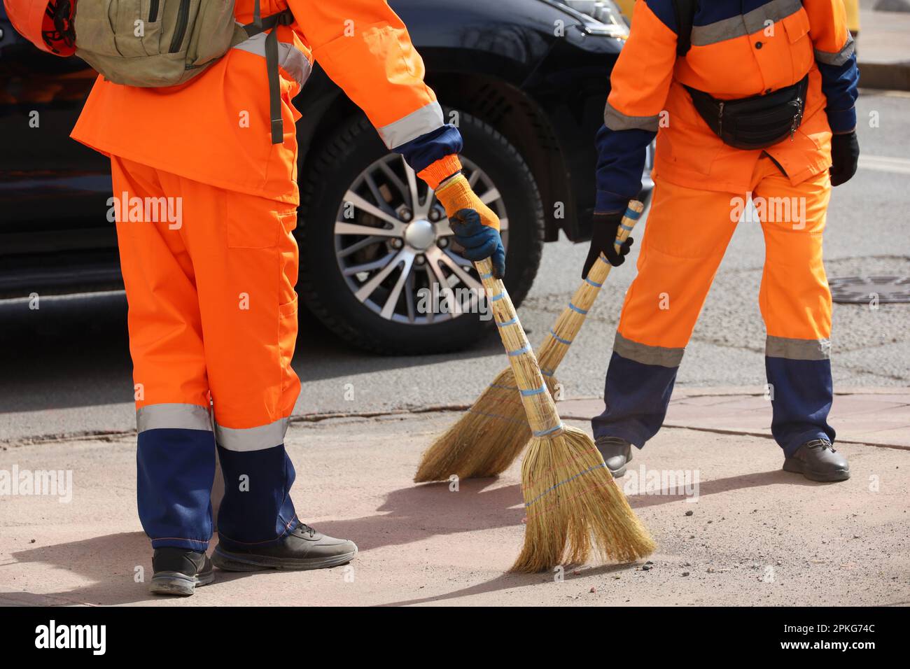 Deux concierges en uniforme orange balayant la rue sur fond de voiture. Nettoyage de la ville de printemps Banque D'Images