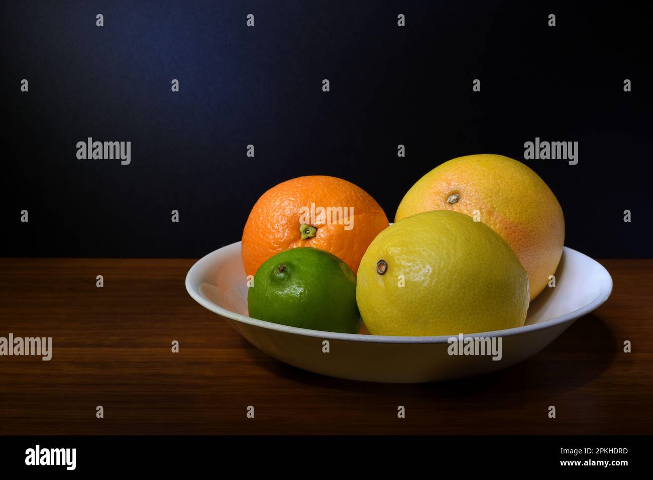 Un bol blanc avec un orange entier, frais, mûr, pamplemousse, citron vert et citron dans un éclairage doux d'ambiance sombre avec un espace de copie à gauche ; capturé dans un studio Banque D'Images