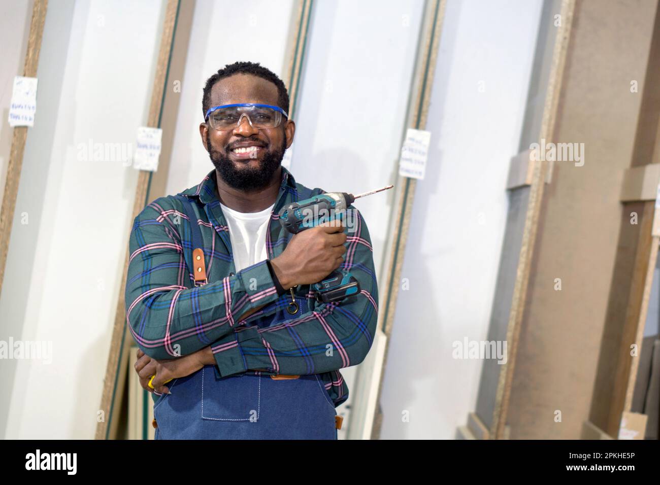 Petit homme à cheveux noirs avec moustache et support à barbe, bras replié, tenant la perceuse électrique sans fil. Le compartiment de rangement en contreplaqué se trouve sur le fond Banque D'Images