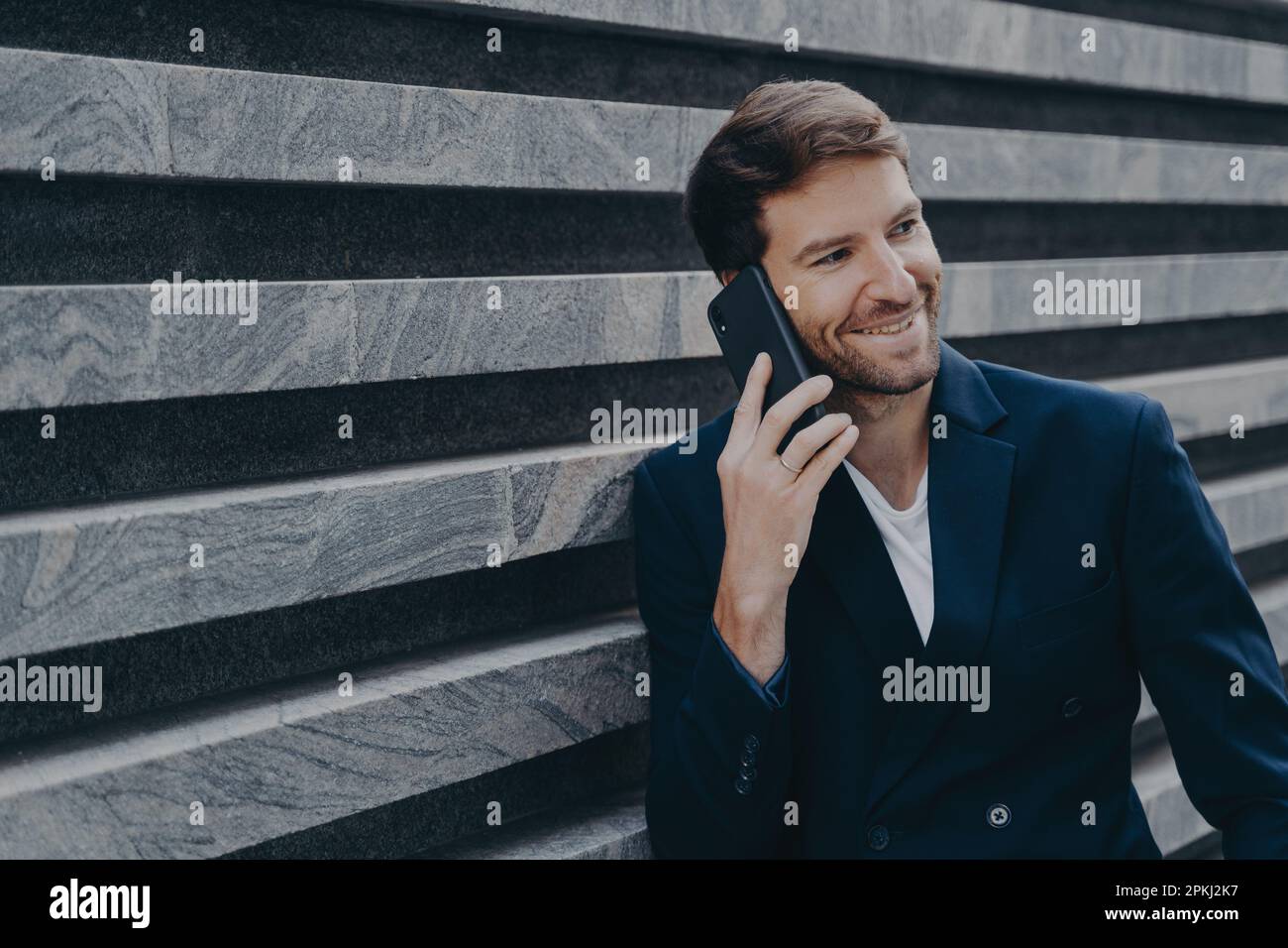 Jeune employé de bureau attrayant dans un blazer élégant parlant sur téléphone mobile pendant les pauses de travail, souriant sincèrement tout en ayant une belle conversation de famille sur smartp Banque D'Images