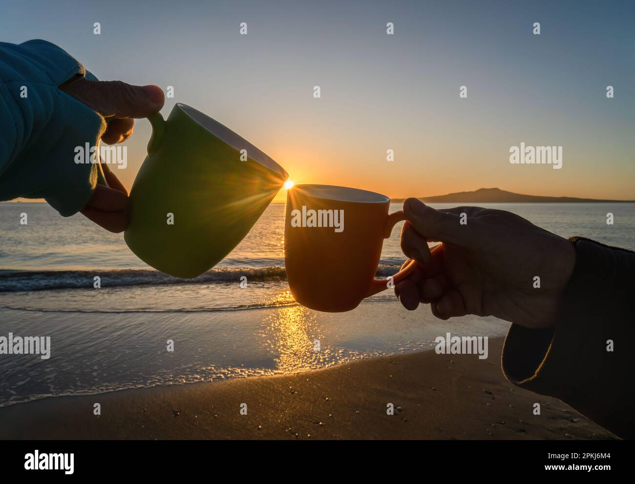 Mains tenant des tasses à la plage de Takapuna au lever du soleil. Le soleil éclate au bord de la tasse. Rangitoto Island au loin. Camping sur la plage Banque D'Images