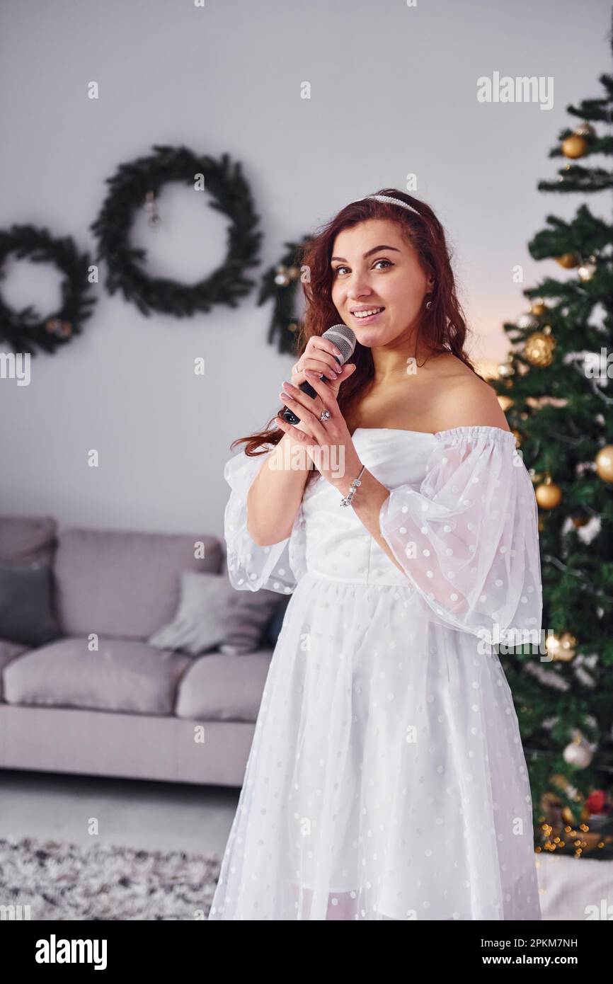 Se tenant dans la chambre intérieure décorée de noël. Femme en robe blanche et avec microphone dans les mains chante dans le . Banque D'Images