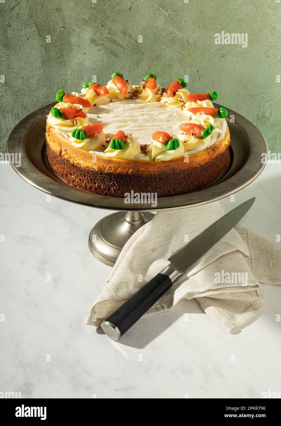 Cheesecake à la carotte pleine plaquée sur un support à gâteau en étain, un couteau et une serviette en tissu au premier plan, sur fond vert. Banque D'Images