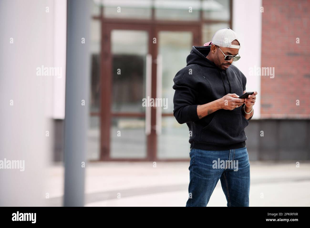 L'homme noir afro-américain utilise un téléphone portable, se tient dans la rue, porte un sweat à capuche, une casquette de baseball et des lunettes de soleil. Portrait modèle mâle élégant regarde h Banque D'Images