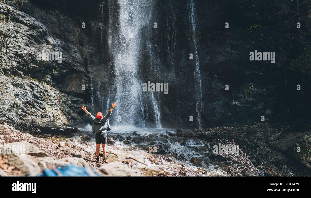 Une femme vêtue de vêtements de randonnée actifs debout près de la cascade de la rivière de montagne s'est levée les bras et appréciant la puissance de la nature éclabousse. Sac à dos sur FO Banque D'Images