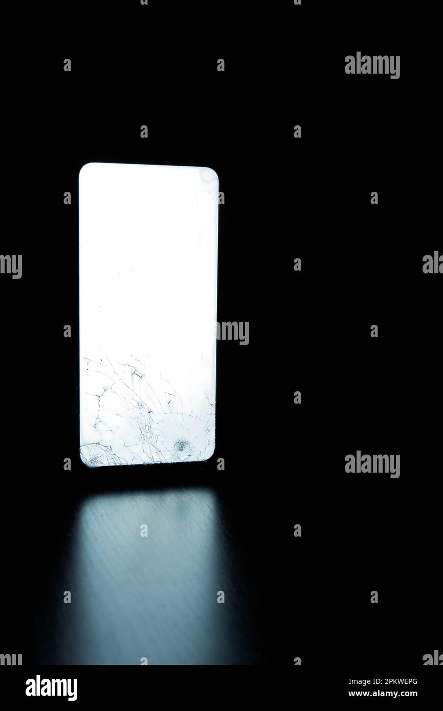 Téléphone portable avec verre brisé et écran blanc sur une surface en bois dans l'obscurité Banque D'Images