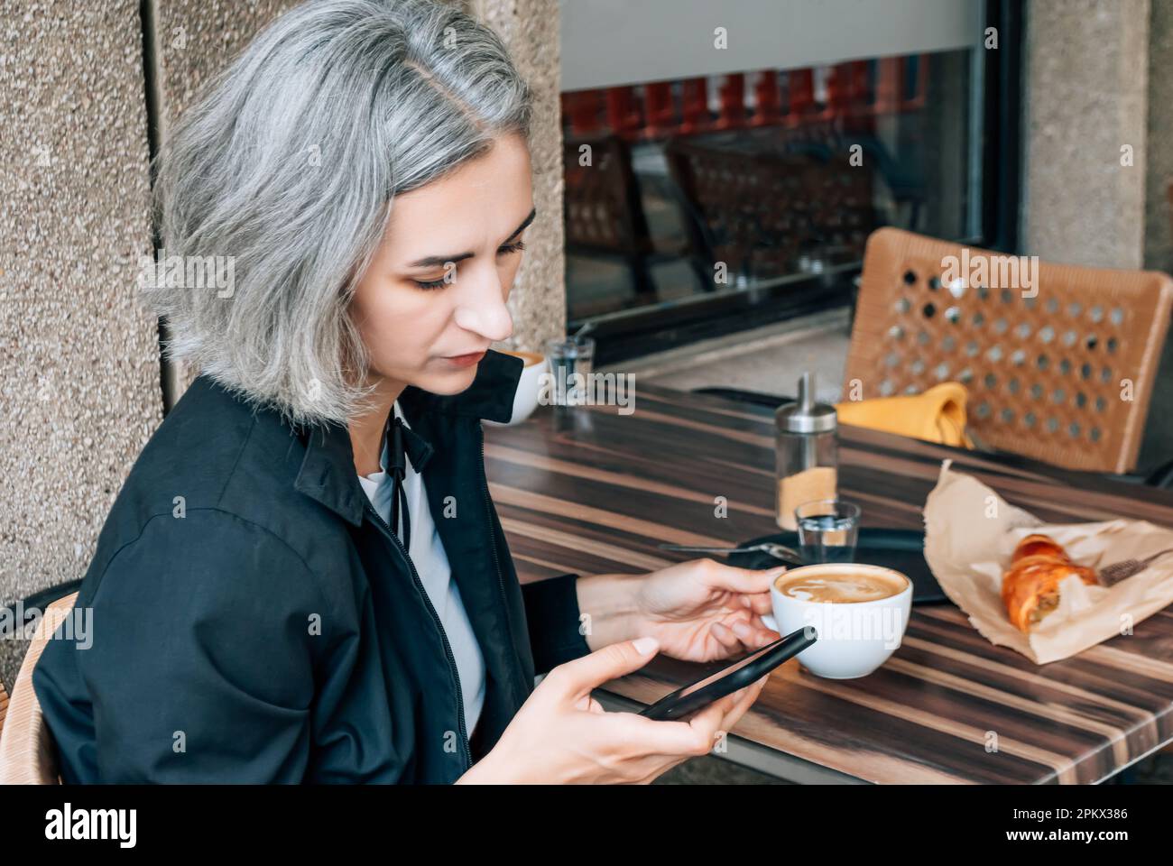 Une femme aux cheveux gris est assise au café de la rue et utilise un smartphone Banque D'Images