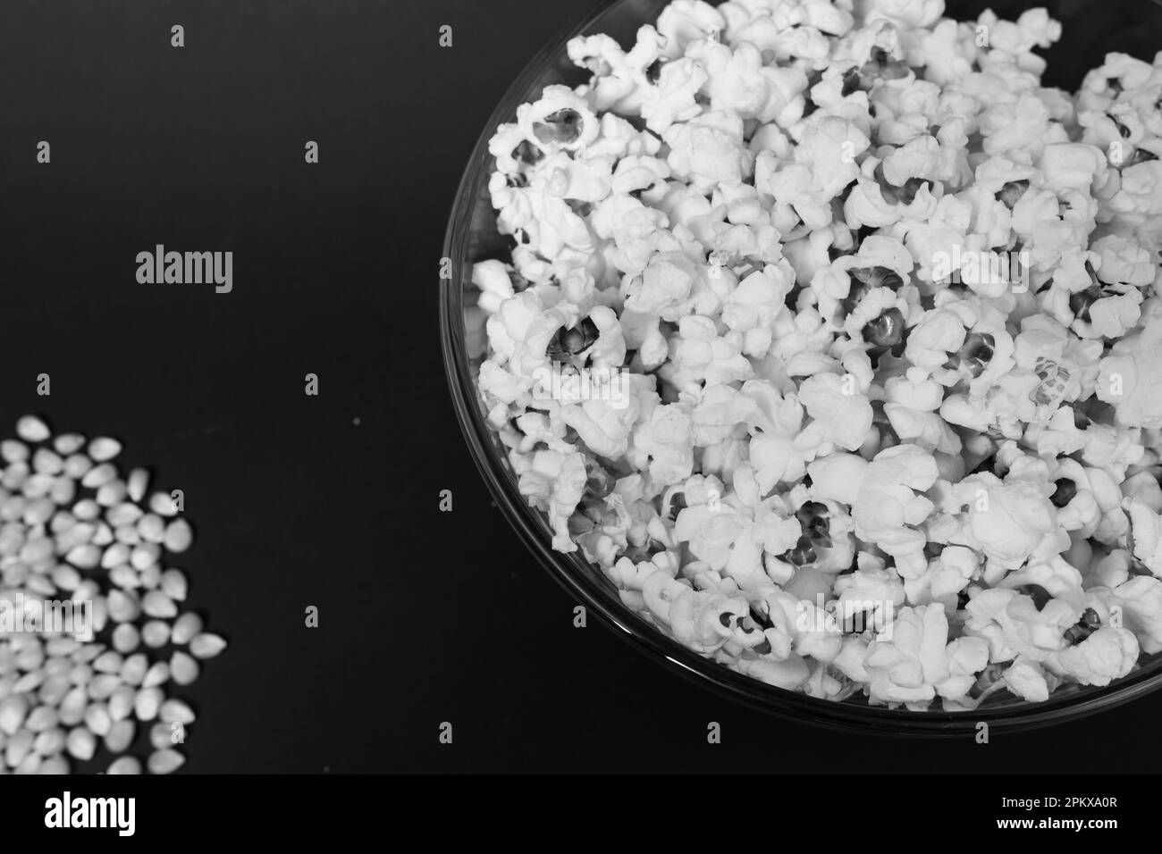 Pop-corn prêt à consommer dans un bol en verre et grains de maïs sur fond noir, vue du dessus et espace de copie. Banque D'Images