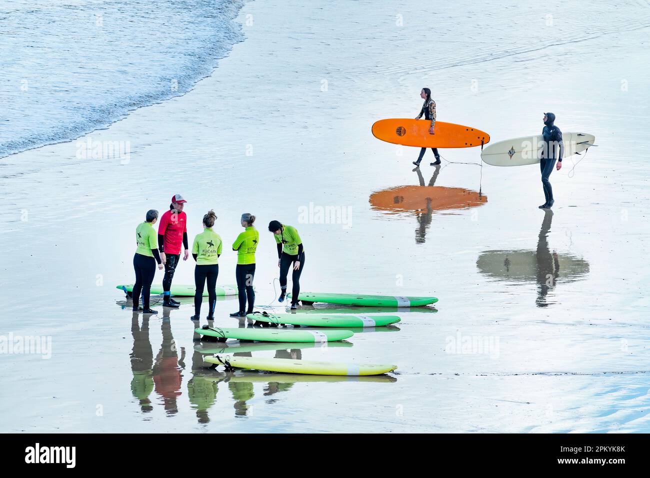 Un groupe d'élèves de sexe féminin instruits à une école de surf à Cornwall. Deux surfeurs expérimentés marchent vers la mer tandis que la leçon continue Banque D'Images