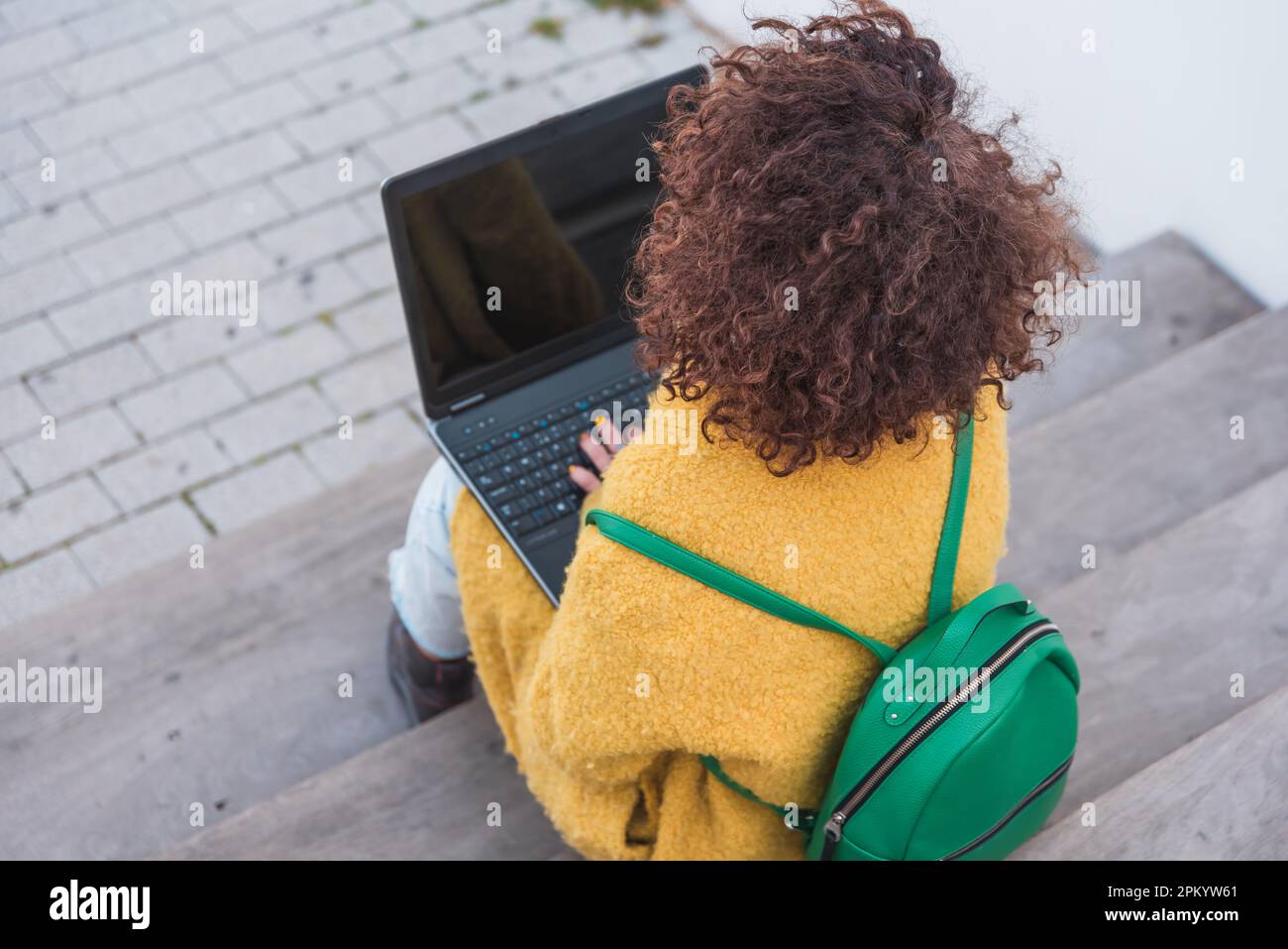 Par-dessus une femme anonyme aux cheveux bouclés dans des vêtements lumineux avec sac à dos assis sur les escaliers et de parcourir l'ordinateur portable tout en préparant les devoirs Banque D'Images