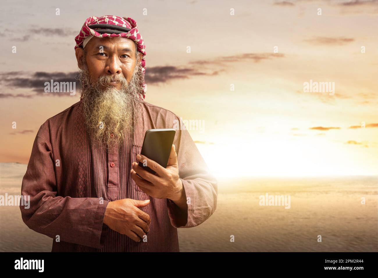 Homme musulman avec une barbe portant du keffiyeh avec agal utilisant un téléphone portable avec le fond de scène de coucher de soleil Banque D'Images