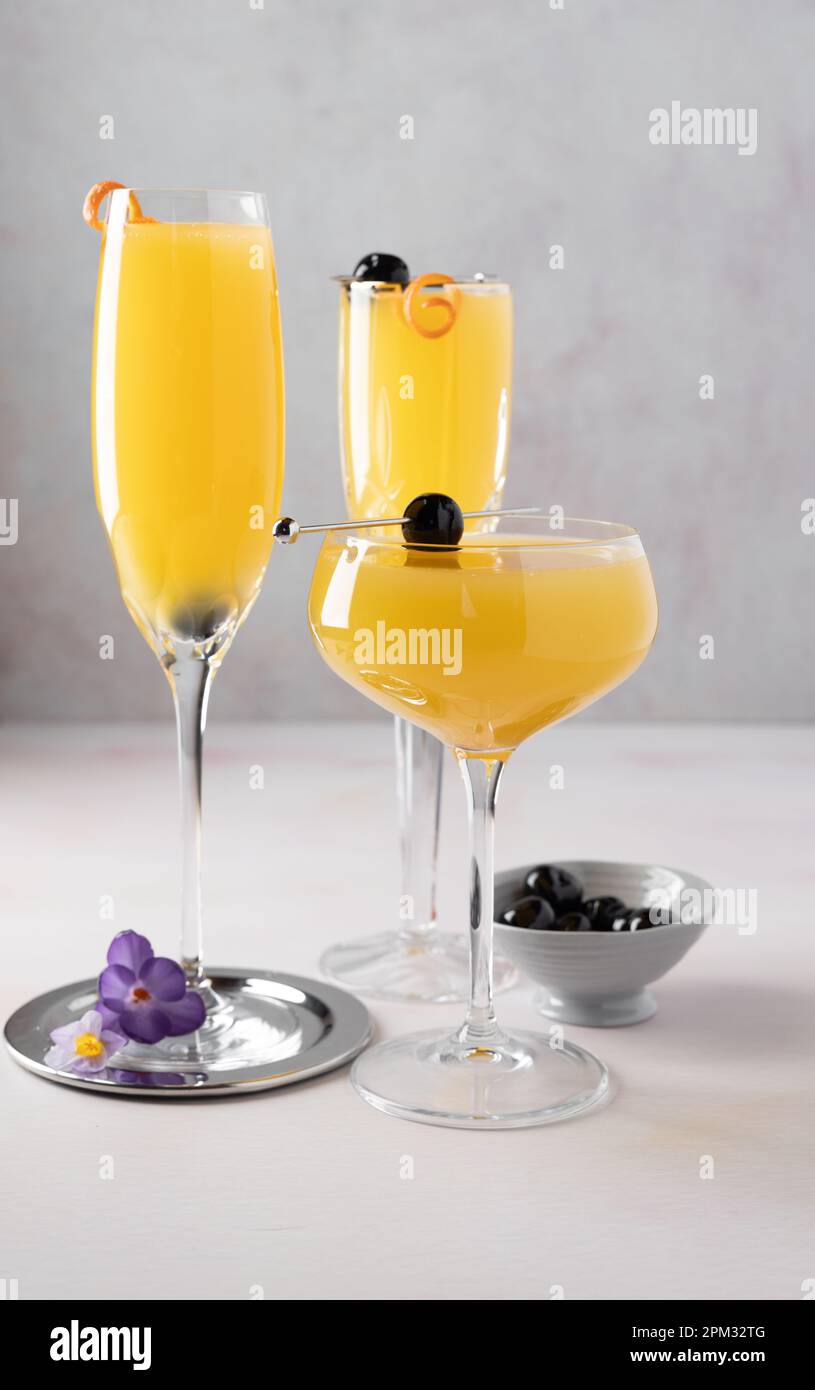 Trois cocktails mimosa en deux flûtes et un verre coupé, des garnitures en cerisier et orange, un bol de cerises, des cocktails et des fleurs de crocus. Banque D'Images