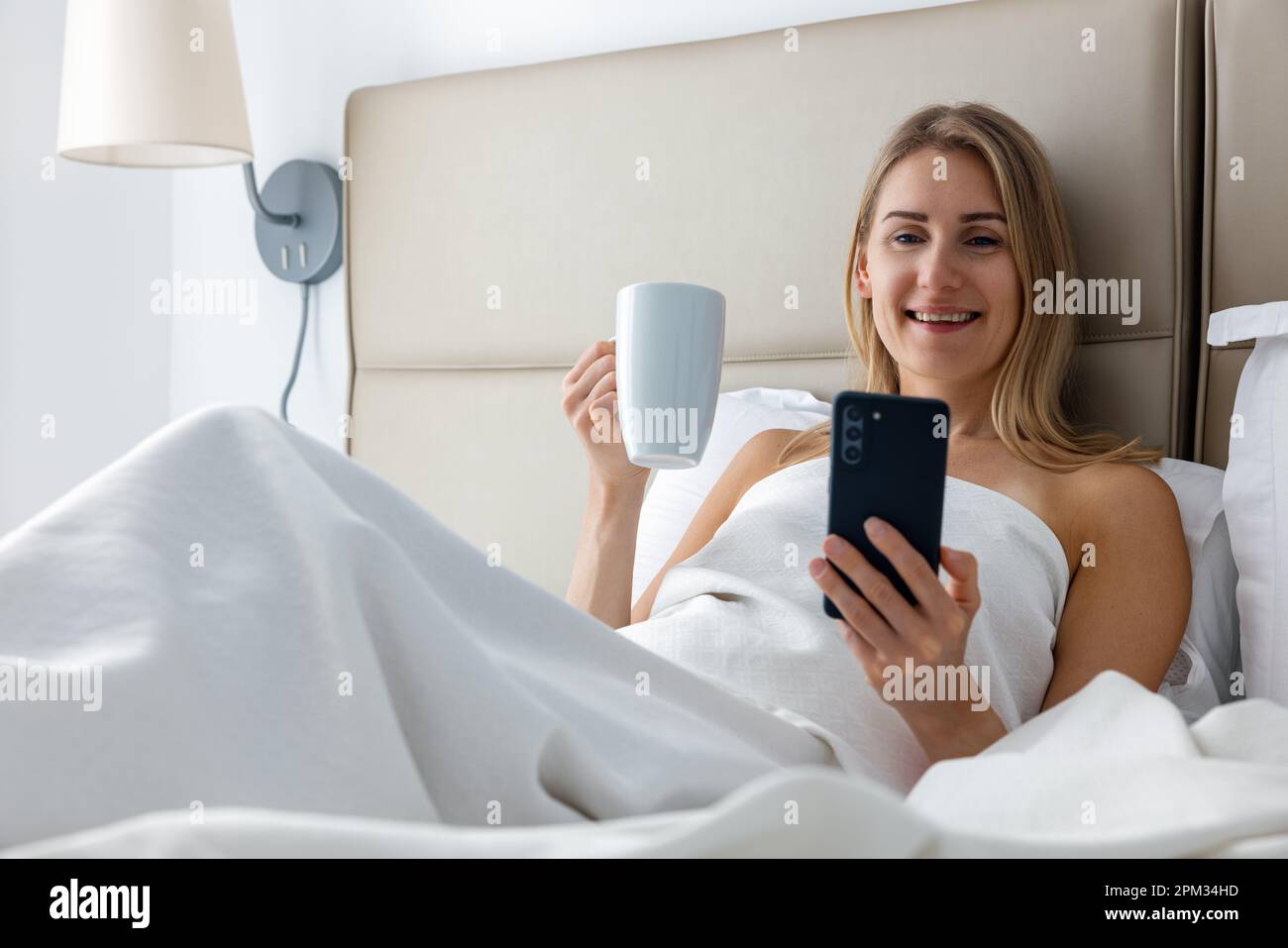 une jeune femme souriante se couche au lit et utilise le téléphone tout en buvant un café le matin dans la chambre à la maison Banque D'Images