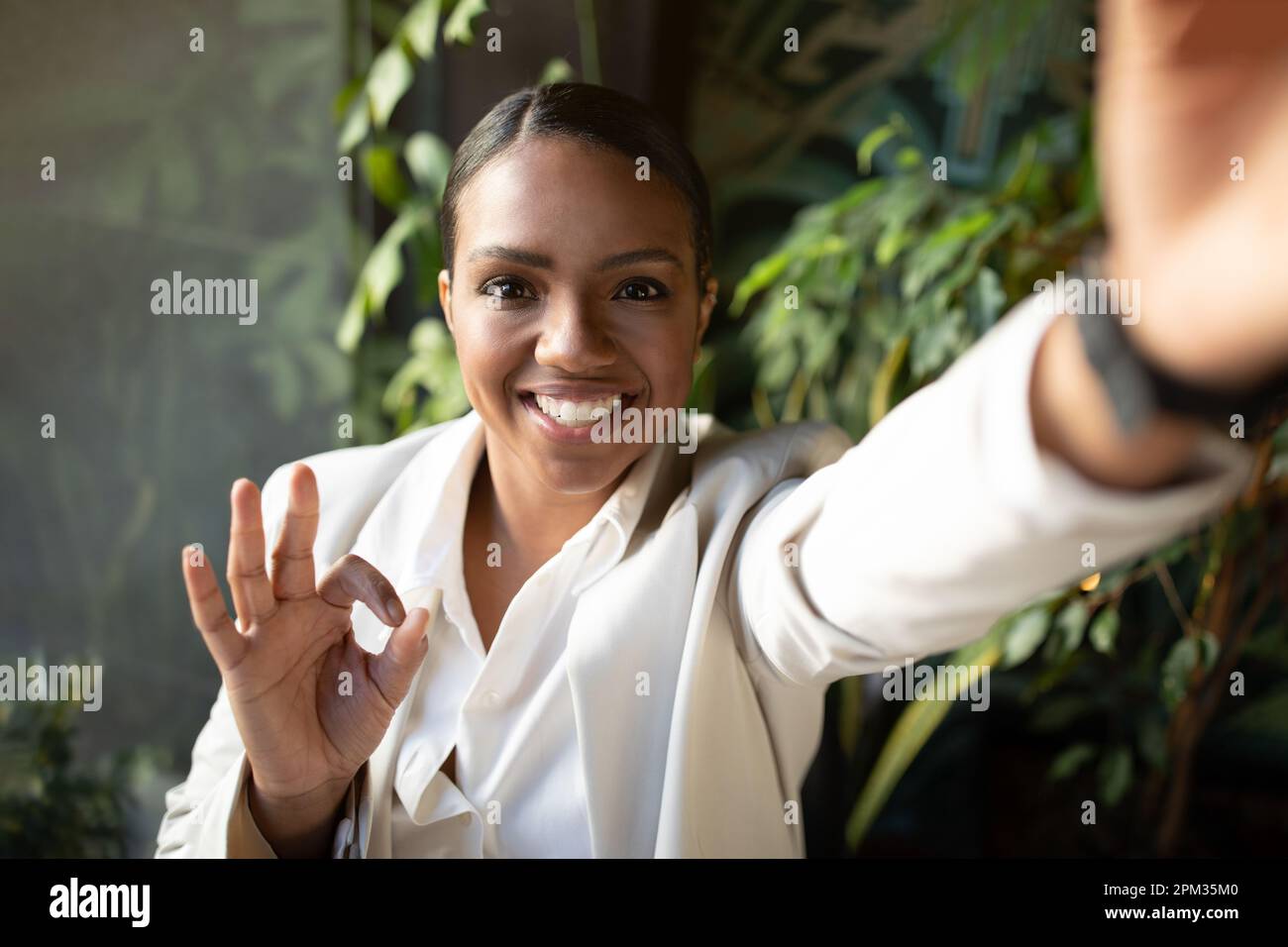 Femme afro-américaine positive du millénaire en costume blanc faisant un geste de main ok, prenant selfie dans le café Banque D'Images