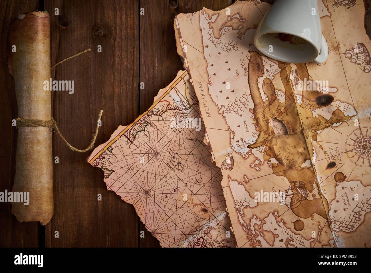 Un vieux parchemin avec de vieilles cartes et une tasse de café renversée sur une table en bois. Banque D'Images