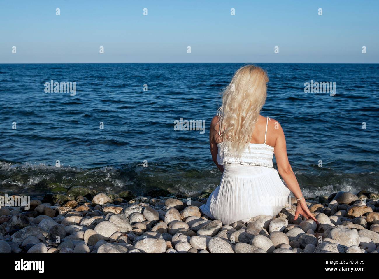 femme blonde en robe blanche, vue de derrière, assise sur une plage de galets, vue sur la mer. Banque D'Images