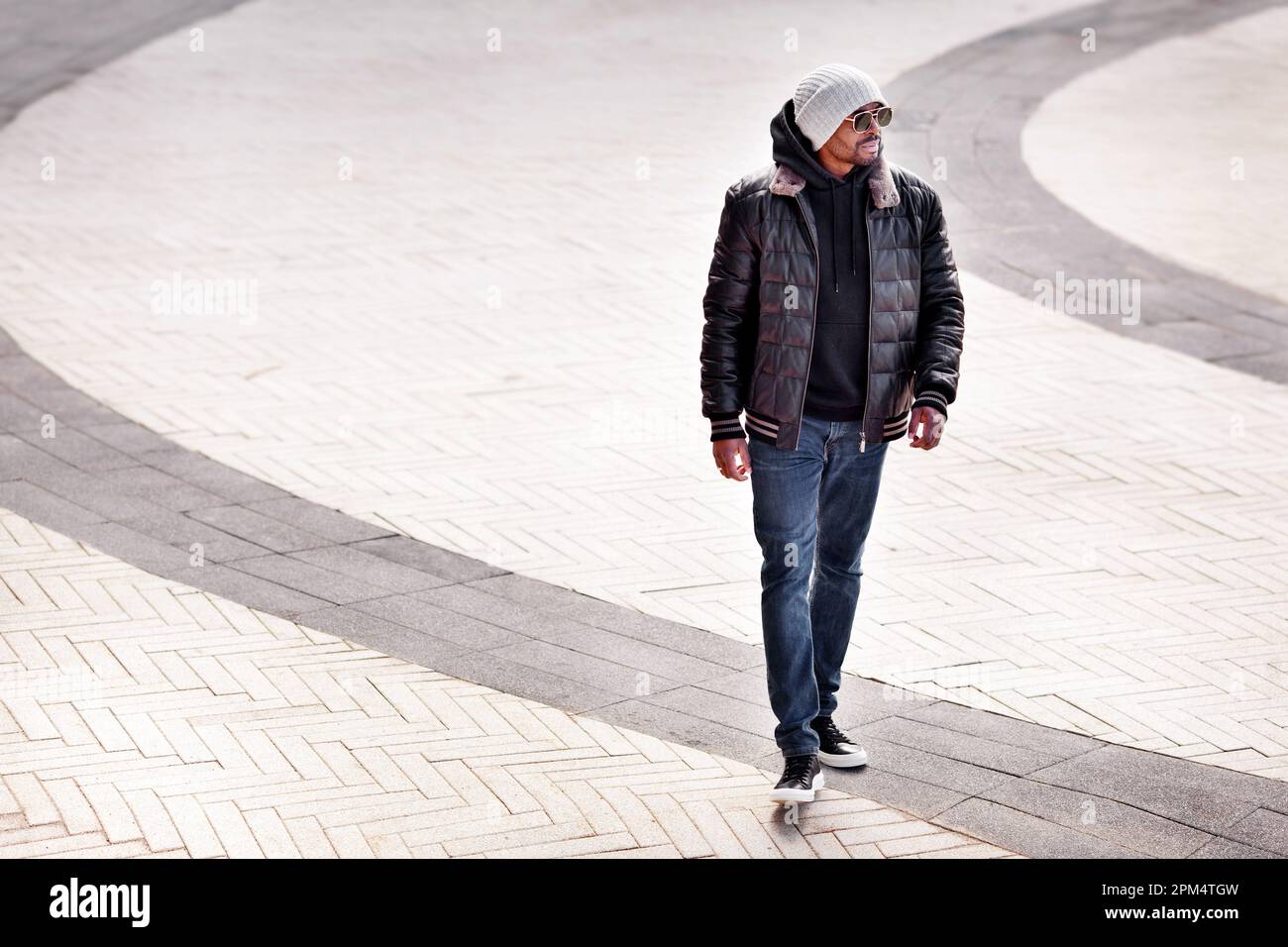 Street fashion, homme afro-américain élégant portant une veste en cuir noir, un sweat à capuche, des baskets, un Jean, un bonnet et des lunettes de soleil marchant sur le dos gris de la rue Banque D'Images