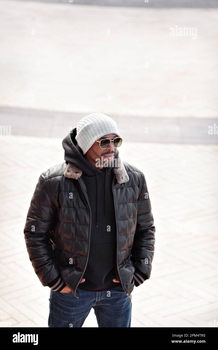 Street Fashion, portrait élégant homme en cuir noir veste, sweat à capuche, bonnet tricoté et lunettes de soleil debout, mains dans les poches Banque D'Images