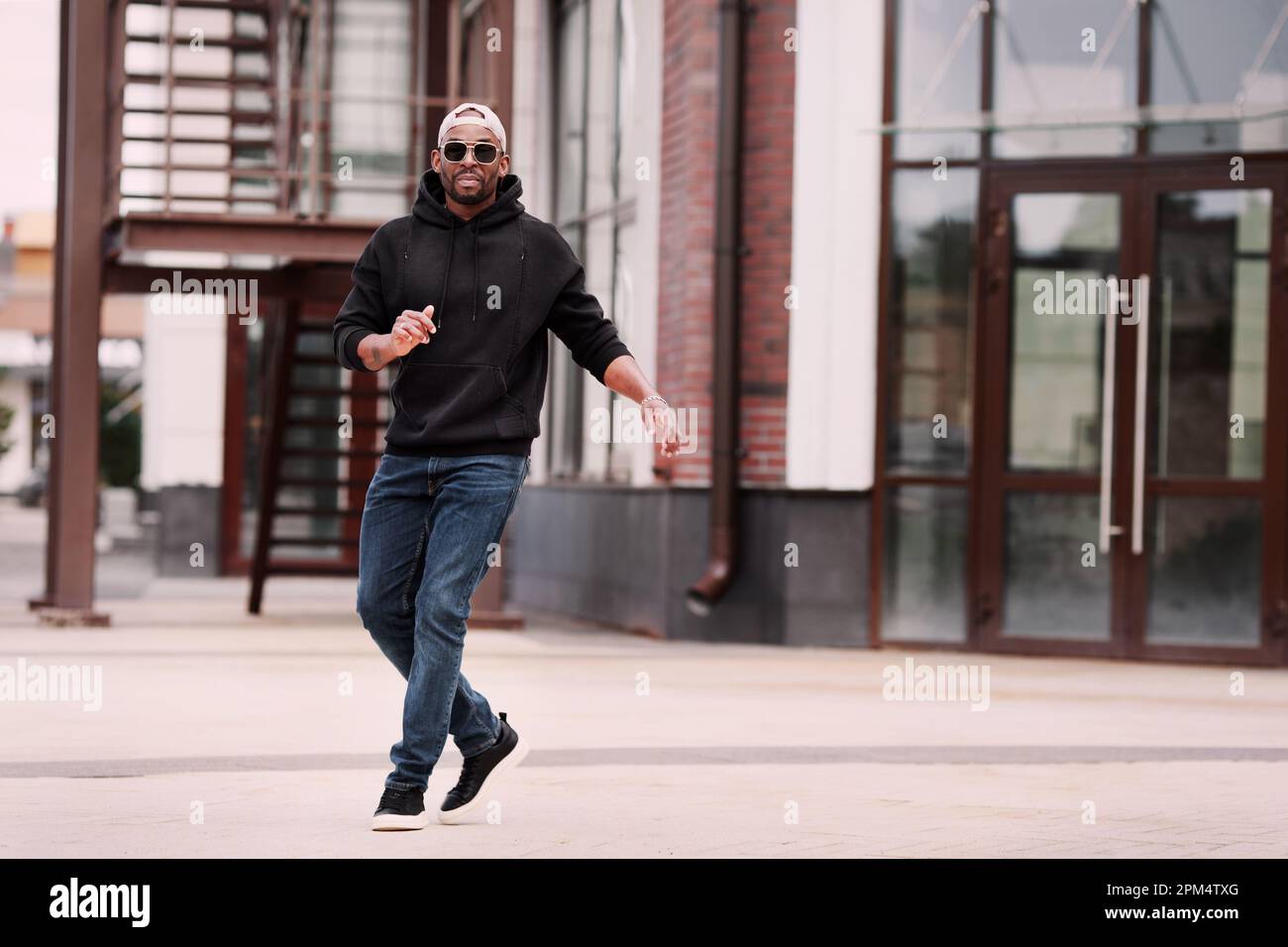 Cool Black man dansant dans la rue de la ville seule, porte noir sweat à capuche, jeans et baskets, casquette de baseball et lunettes de soleil Banque D'Images