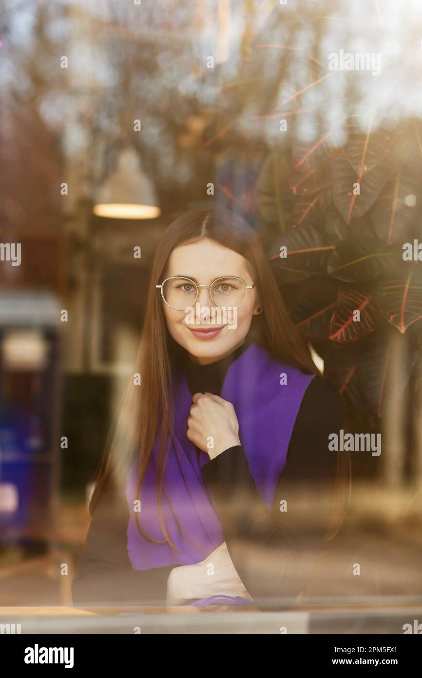 une jeune femme en lunettes et un foulard violet derrière une fenêtre de café Banque D'Images