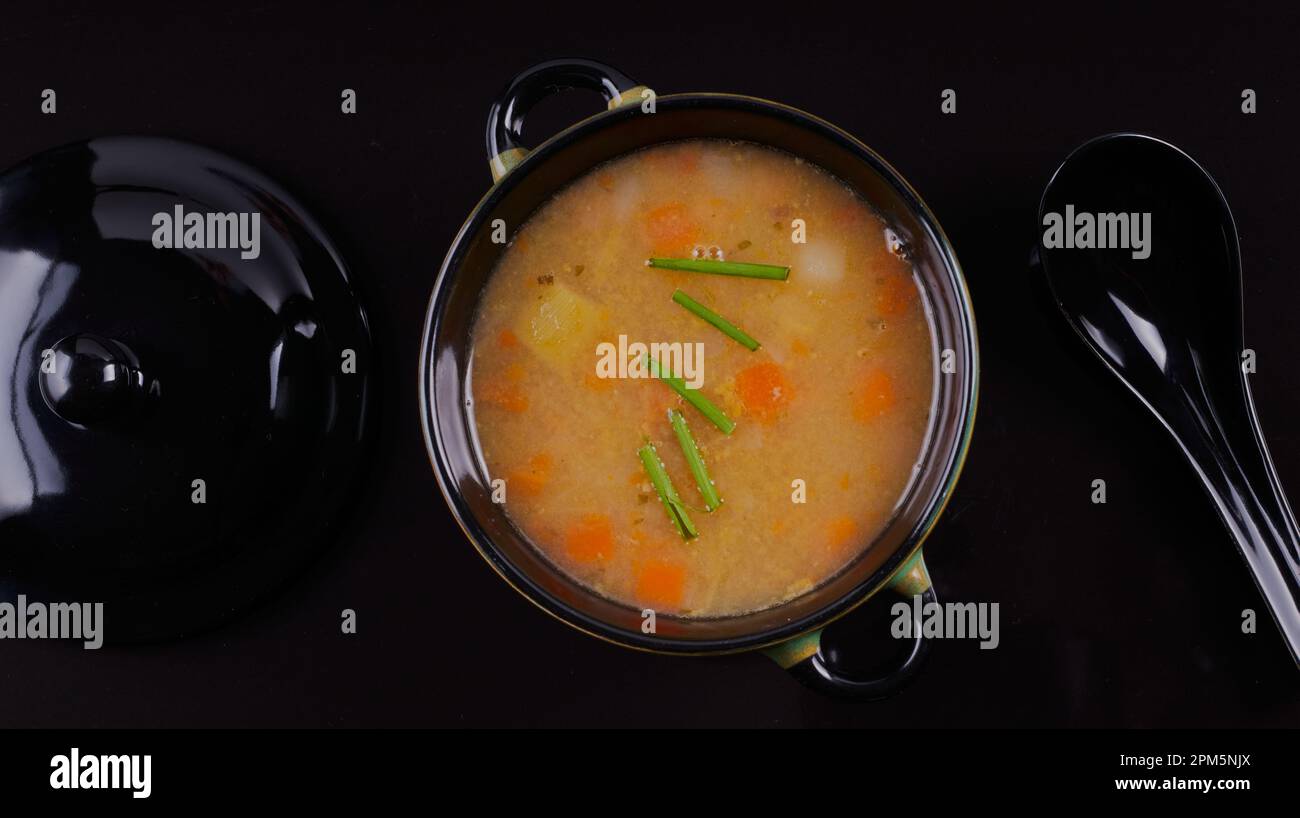 Bouillon de légumes maison dans un bol noir avec cuillère noire sur une surface noire. Banque D'Images