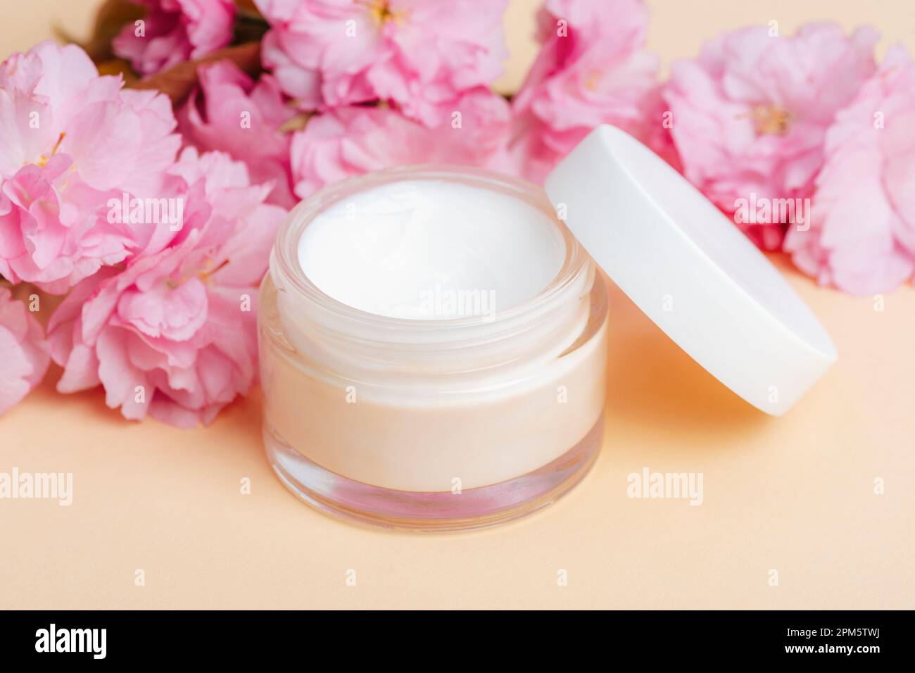 Crème visage dans un pot et fleur de cerisier rose sur fond beige. Soin de la peau, concept de traitement de beauté. Gros plan. Banque D'Images