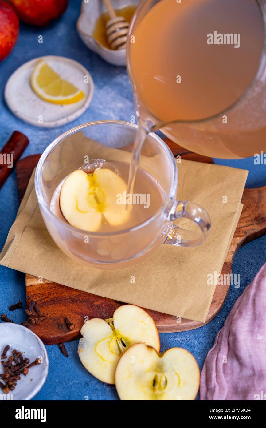 Thé de pomme étant versé dans une tasse de verre, des tranches de pomme, des clous de girofle, des tranches de citron, du miel autour de lui. Banque D'Images