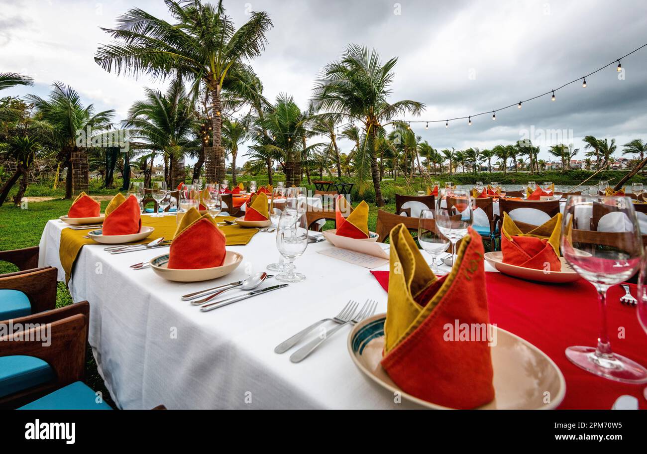 Plusieurs longues tables à dîner dehors dans un cadre tropical avec de nombreux palmiers et des serviettes rouges avec couverts. Banque D'Images