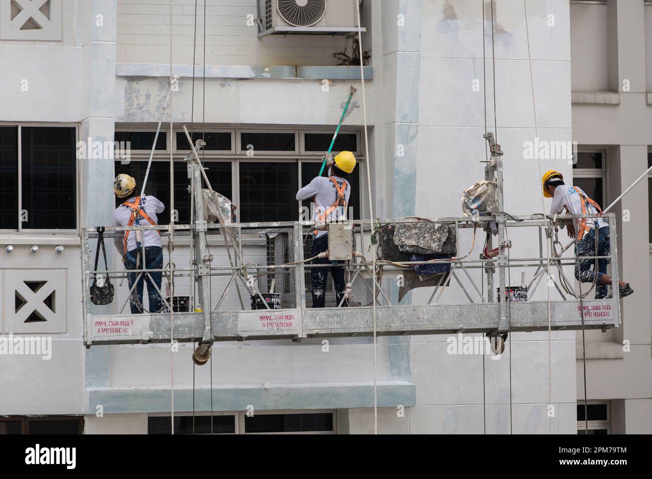 Trois ouvriers sur les ascenseurs échafaudés en hauteur, travaillant de leur mieux pour mettre au rebut la peinture pelable, enduisant la nouvelle peinture sur la façade du bâtiment. Banque D'Images