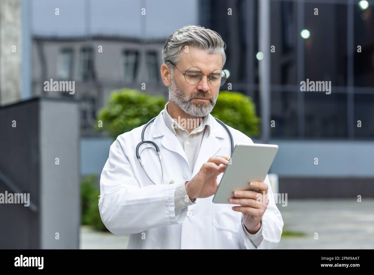 Médecine en ligne. Un médecin de sexe masculin plus âgé aux cheveux gris se tient à l'extérieur de la clinique sous un manteau blanc et utilise un comprimé. Numérotations, appels, SMS, discussions. Banque D'Images