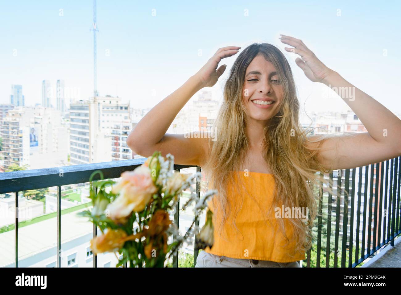 portrait d'une jeune femme latine d'origine colombienne, vêtue de jaune, assise sur son balcon d'appartement, confondue avec les mains sur la tête, souriant et lo Banque D'Images