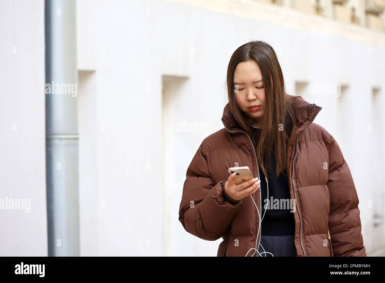 Fille asiatique dans un casque portant une veste marron marchant avec un smartphone dans une rue. Utilisation d'un téléphone portable au printemps Banque D'Images