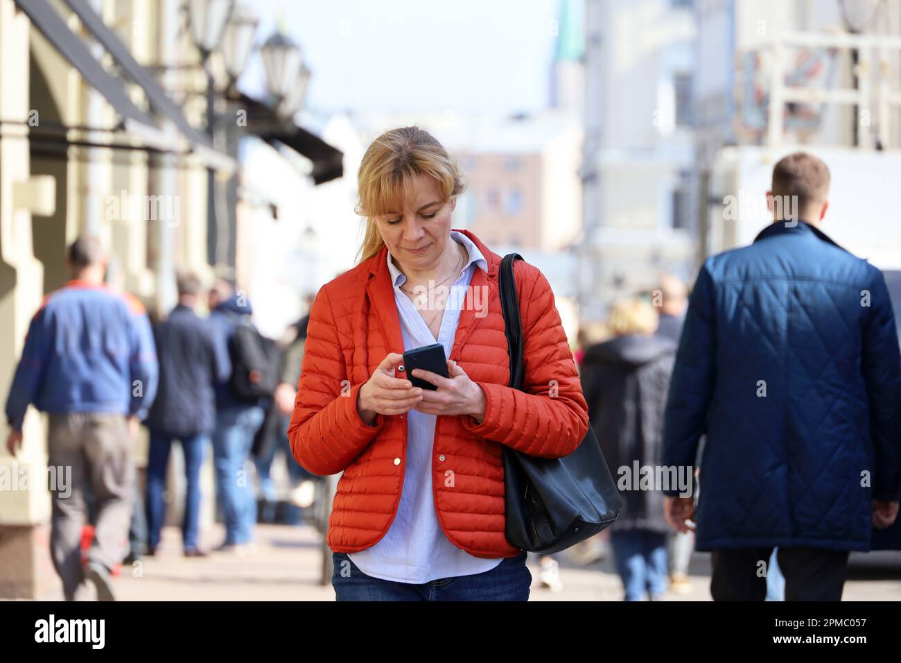 Femme en veste rouge marchant dans une rue avec un smartphone sur le fond des gens. Utilisation d'un téléphone portable au printemps Banque D'Images