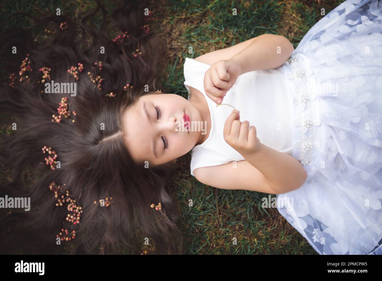 Petite fille en robe blanche couchée sur l'herbe, elle sourit, ses cheveux sont très longs et princesse-comme, elle joue avec ses mains sur le thème de l'enfant Banque D'Images