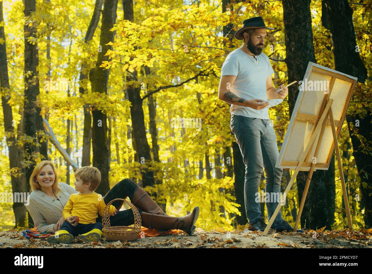 Beau barbu homme dessine une image de sa jeune famille dans le parc à l'aide d'une palette de peintures et d'une spatule. Chevalet de dessin et toile avec un Banque D'Images