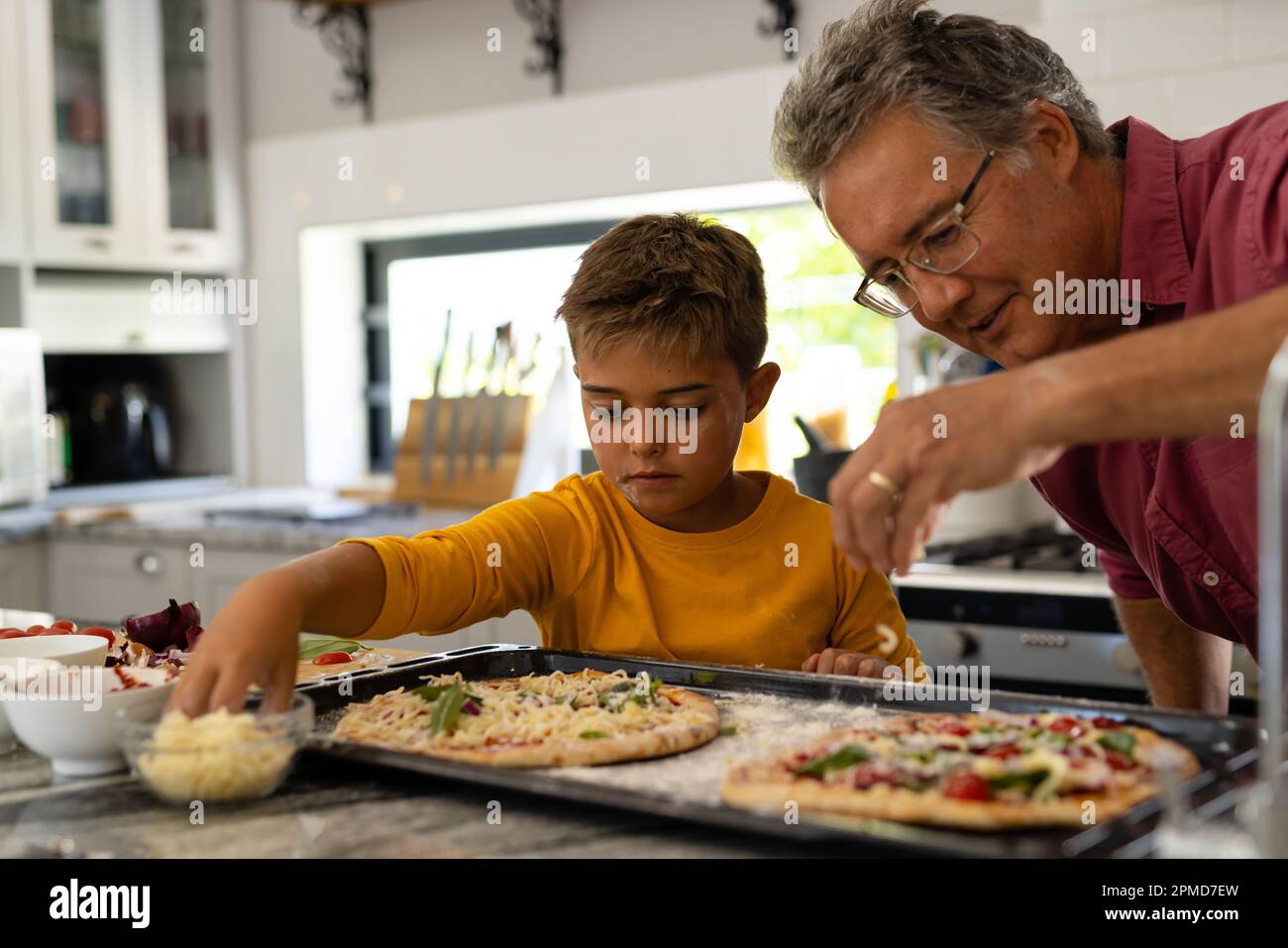 Grand-père et petit-fils caucasiens qui garnissent du fromage sur une pizza au-dessus de l'île de cuisine Banque D'Images
