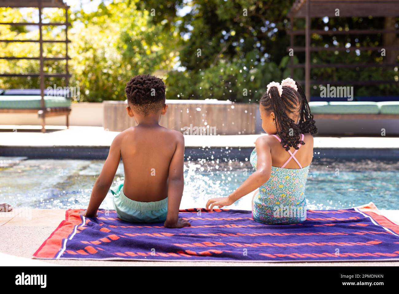 Vue arrière de frères et sœurs afro-américains qui éclabousse de l'eau dans la piscine tout en étant assis sur une serviette au bord de la piscine Banque D'Images