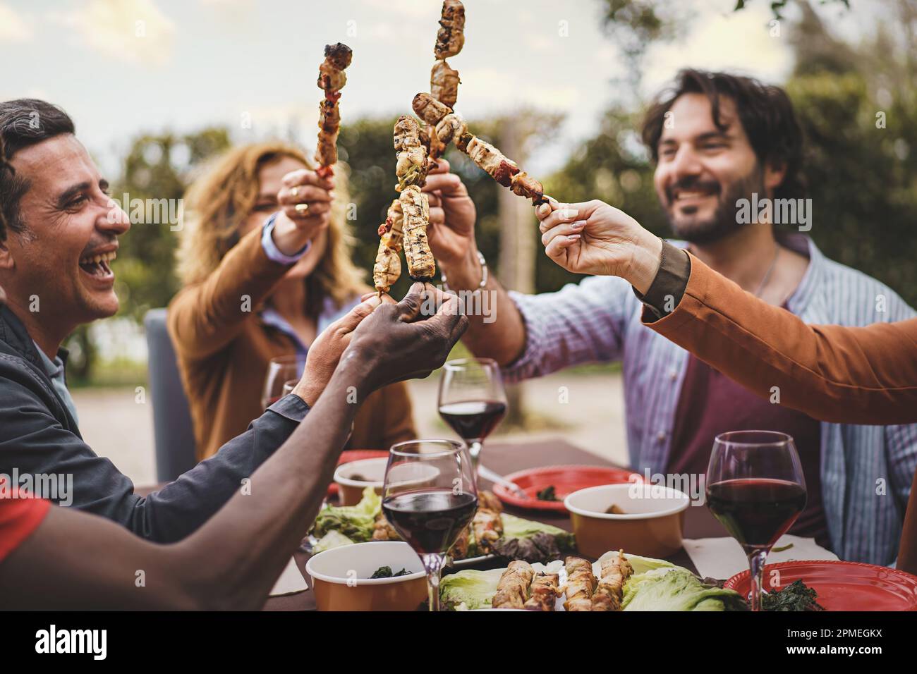 Un groupe de personnes dans leurs 30s et 40s assis à une table de jardin, tenant des brochettes de viande et de toaster. Concentrez-vous sur les brochettes. Appartenance ethnique mixte, y compris un yo Banque D'Images