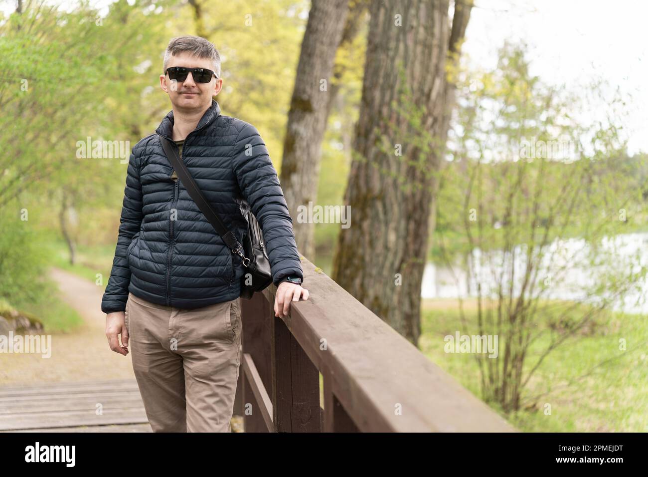 Un homme d'âge moyen en lunettes de soleil et une mince veste en duvet sur une passerelle en bois près du lac au début du printemps Banque D'Images